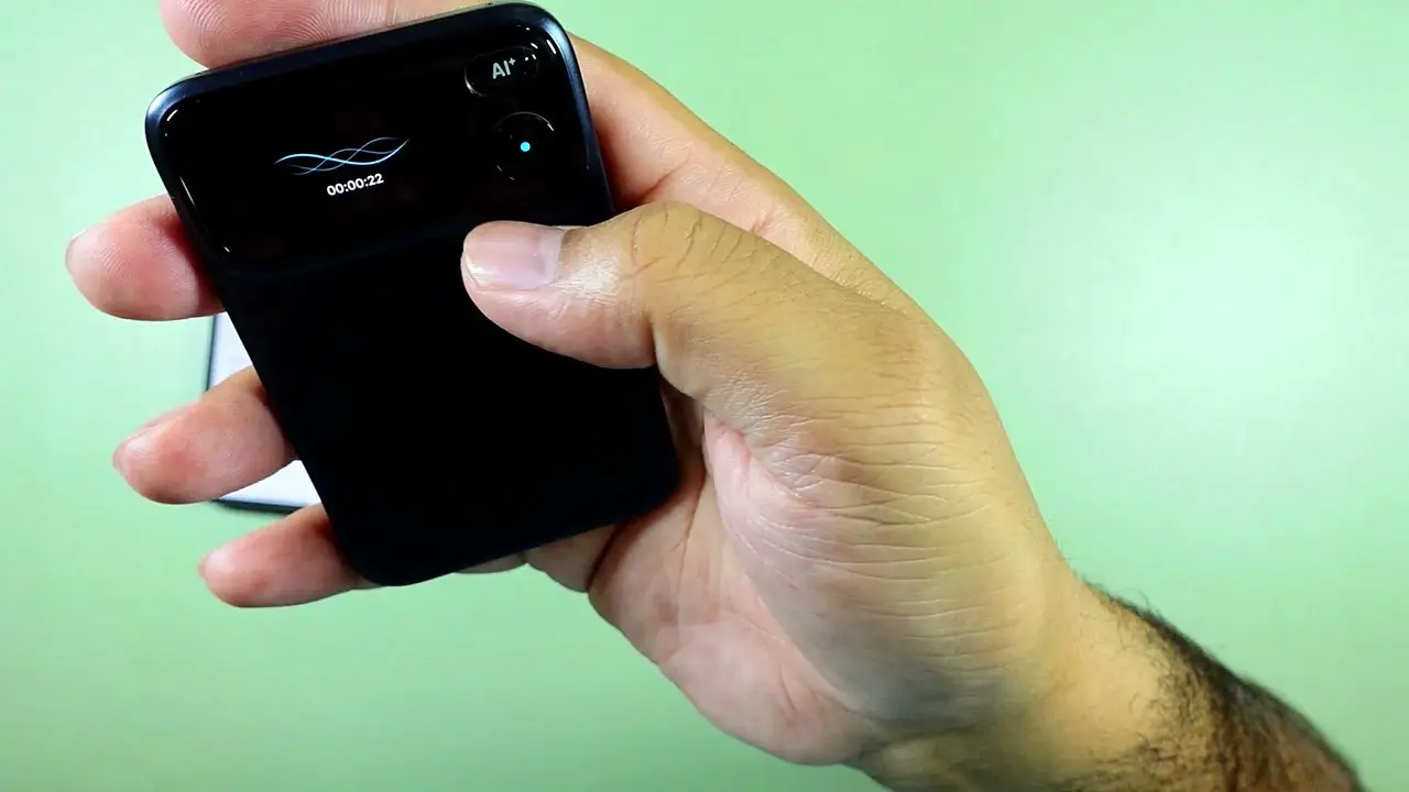 A hand holding the black Comu Action Pro with a waveform, recording timer and cyan button indicator visible