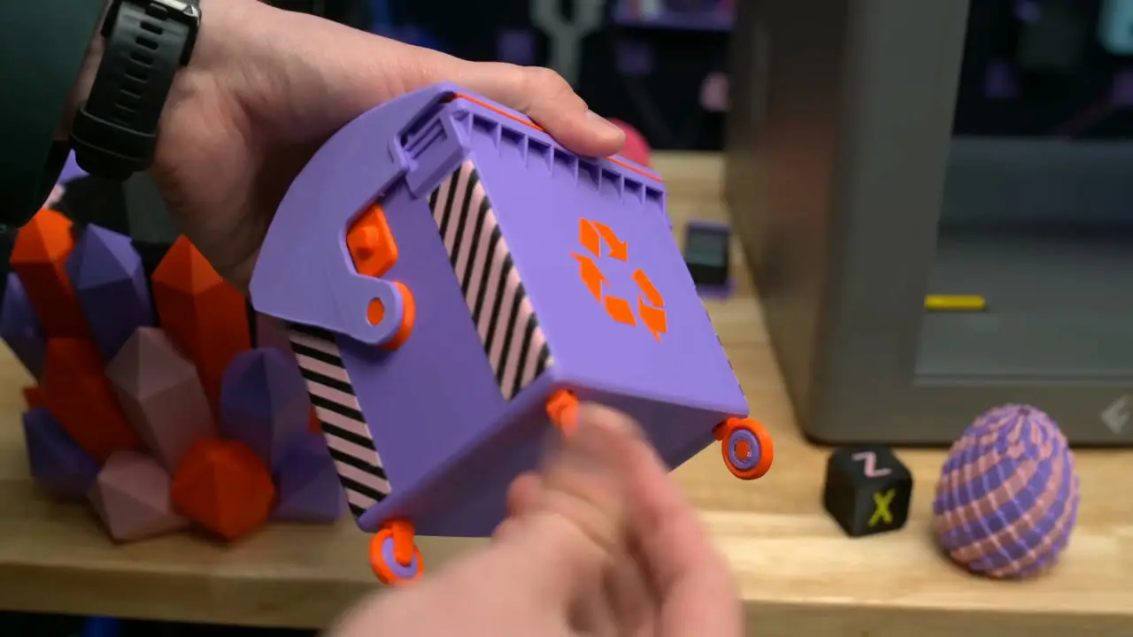 Hands holding a purple and orange printed miniature bin with a hinged lid and wheels