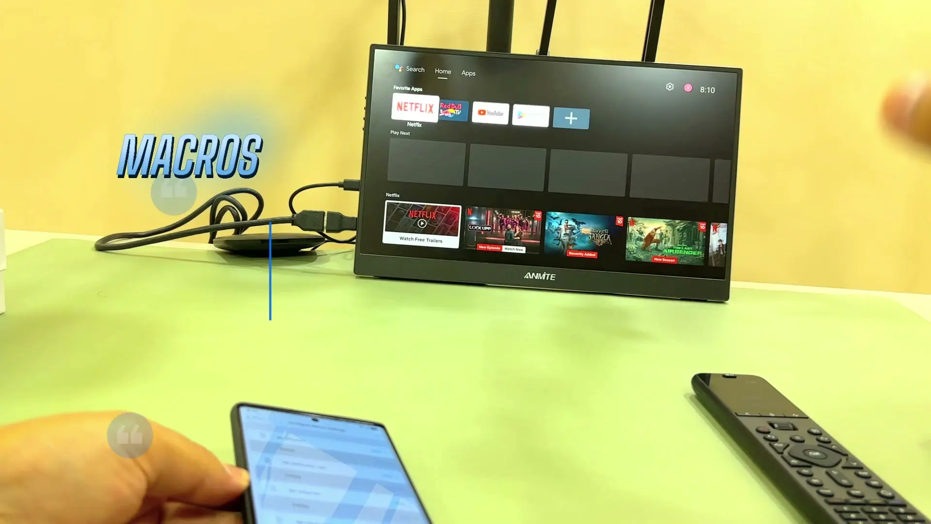 A Google TV home screen with Netflix, Red Bull TV and YouTube app icons, with on-screen text reading MACROS, and the U3 remote and a phone visible in the foreground