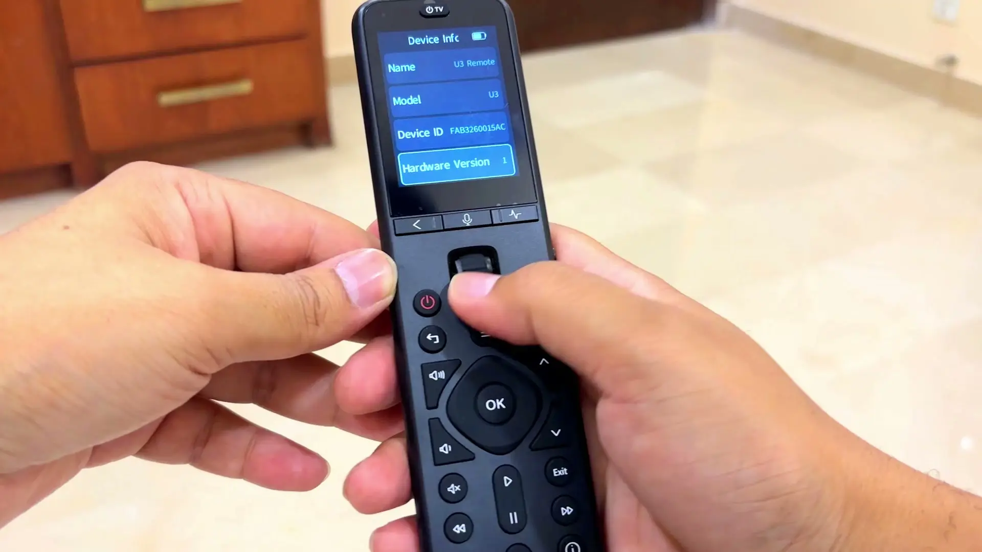 A hand holding the SofaBaton U3 remote with its screen showing a Device Info page listing name, model, device ID and hardware version, thumb resting near the scroll wheel