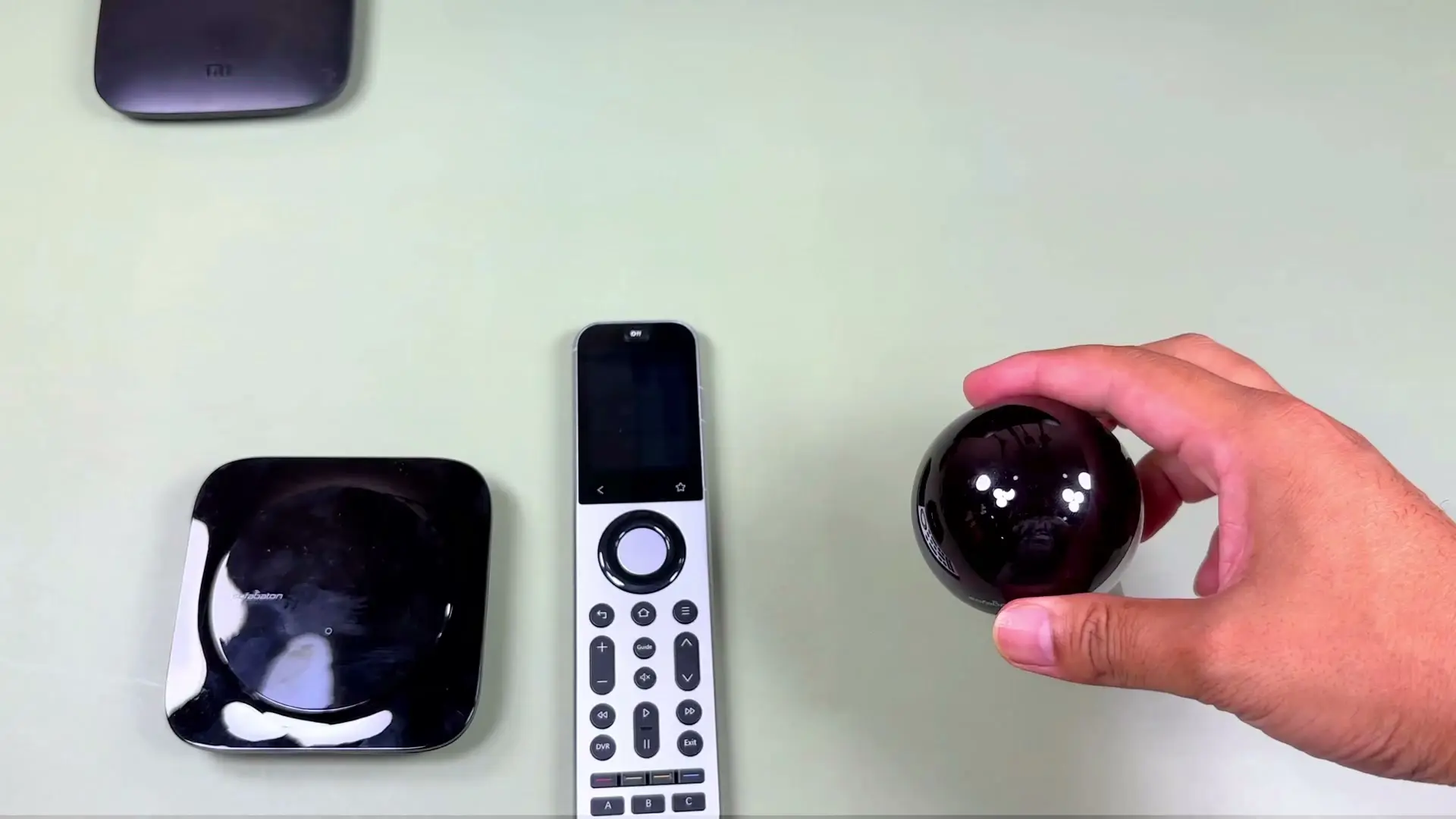The Sofabaton X2 remote seen from above showing its touchscreen and physical buttons — a directional pad, playback, volume and lettered keys — beside the hub and the R1 repeater held in a hand