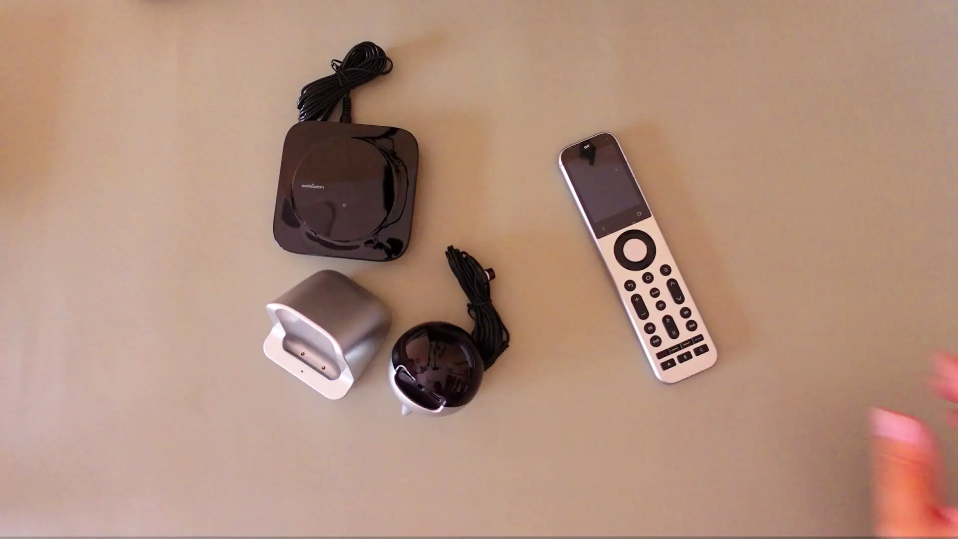 The Sofabaton X2 Set laid out on a tan surface: the glossy black hub, the silver charging dock, the R1 repeater with its coiled cable, and the silver X2 remote with a touchscreen and physical buttons