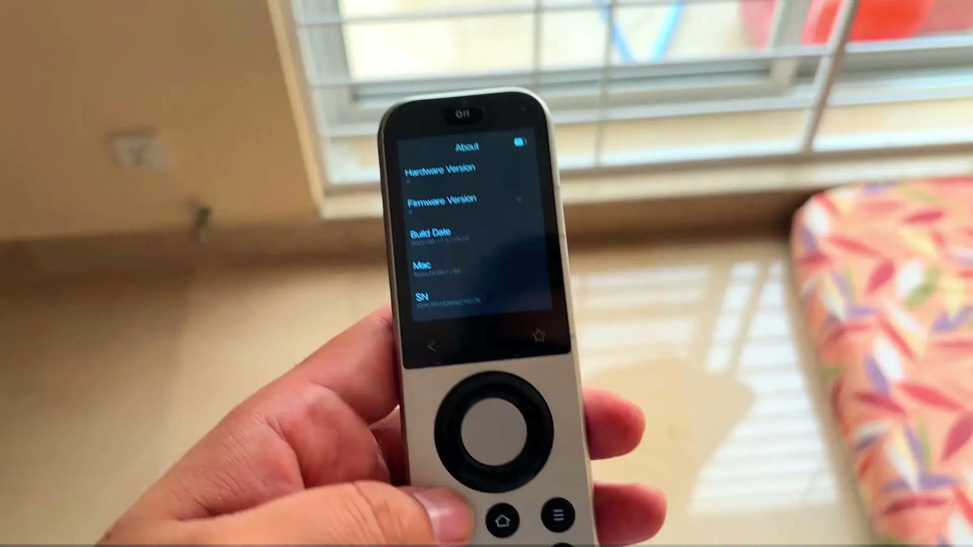 A hand holding the Sofabaton X2 remote, its touchscreen showing an About screen with hardware version, firmware version and build date