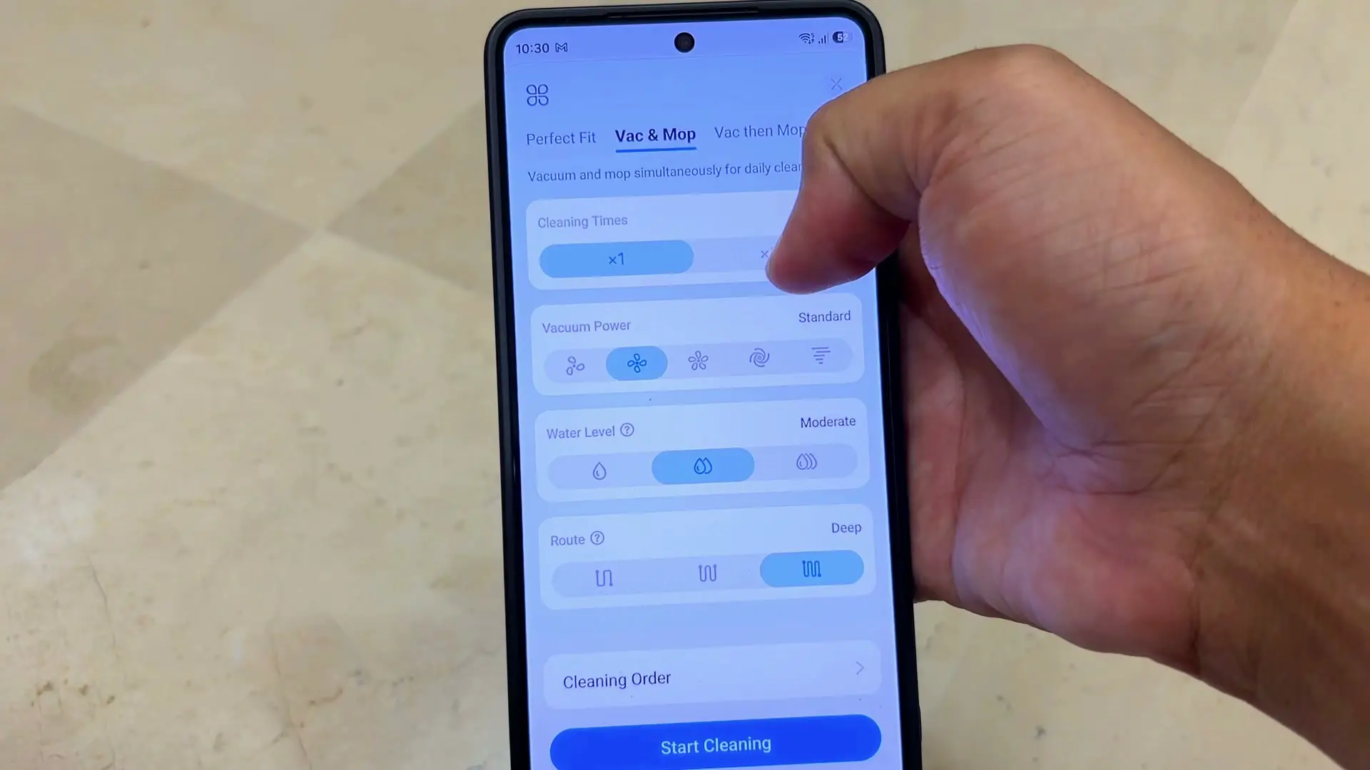 A hand holding a phone showing the Tapo app's Vac and Mop screen with Cleaning Times, Vacuum Power, Water Level and Route options and a Start Cleaning button