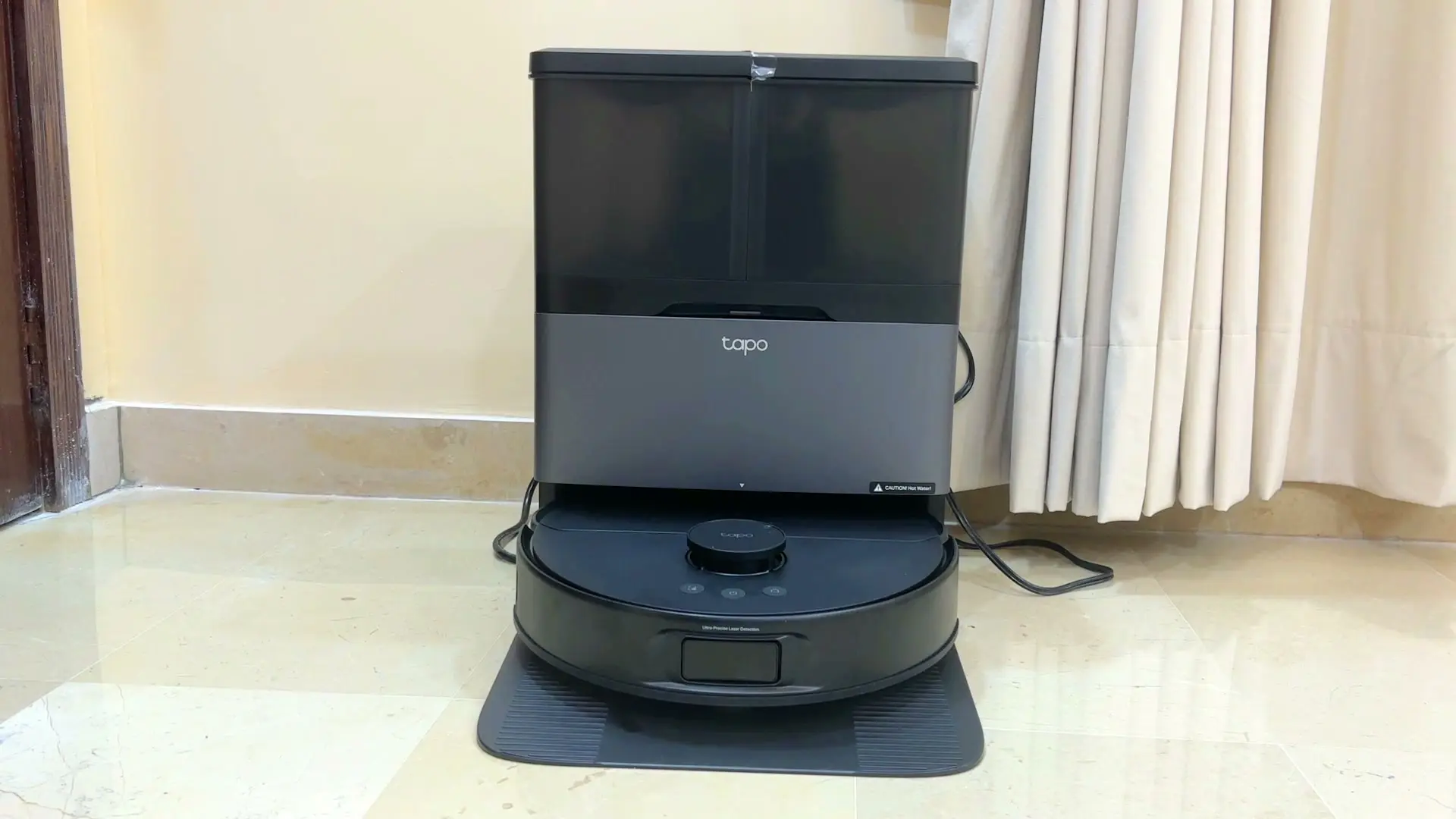 The Tapo RV50 Pro Omni robot vacuum parked in its tall black self-emptying dock on a marble floor, with a 'Caution! Hot Water!' label on the dock and a curtain behind it