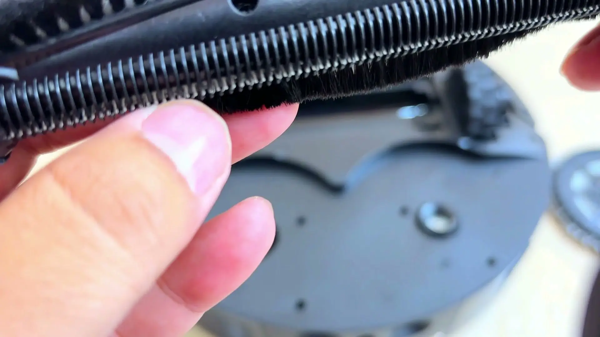 A close-up of the robot's anti-tangle main roller showing a comb of fine teeth and dense bristles, with a finger held against it for scale