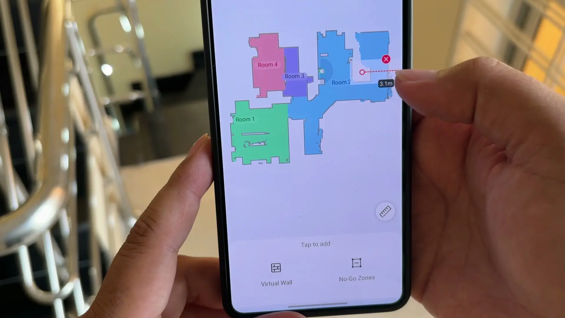 The Tapo app floor map showing four coloured rooms with a 3.1-metre virtual-wall line being drawn across it, Virtual Wall and No-Go Zones buttons below, and a metal stair railing blurred behind