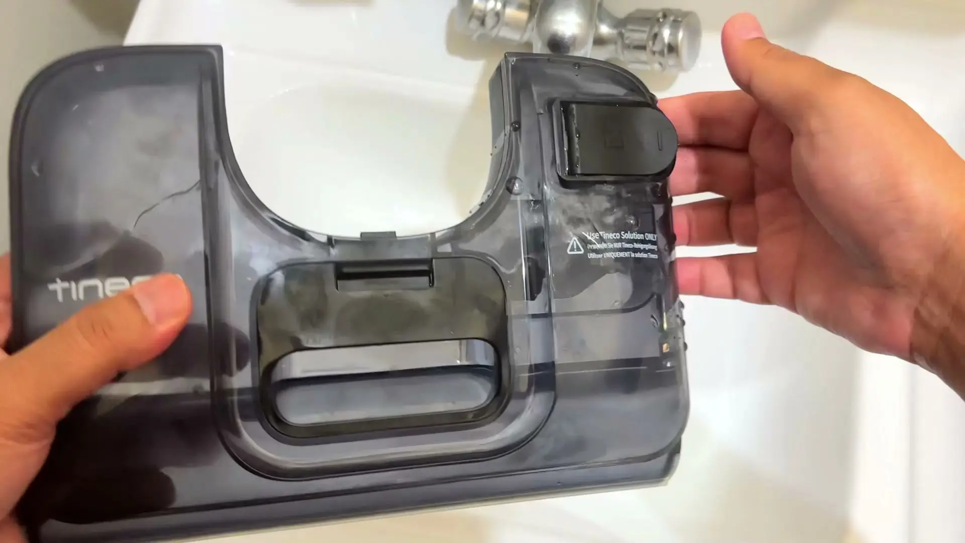 Two hands holding the Tineco's clear dirty-water tank over a sink, with a 'Use Tineco Solution ONLY' label visible on it