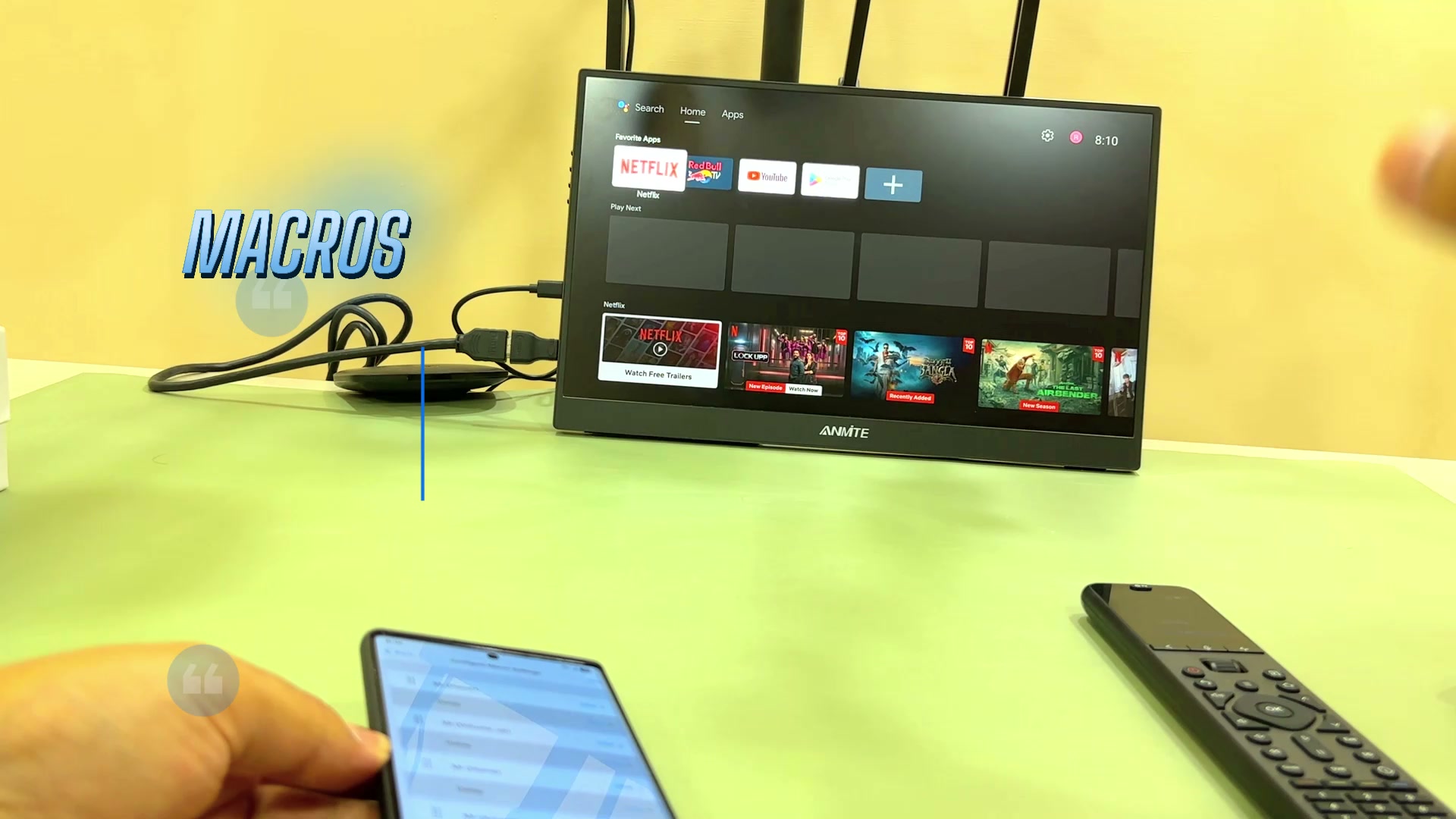 A Google TV home screen with Netflix, Red Bull TV and YouTube app icons, with on-screen text reading MACROS, and the U3 remote and a phone visible in the foreground